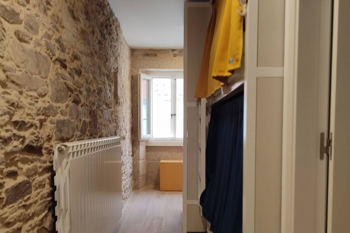 A narrow hallway with a stone wall on one side and a white radiator on the other. There is a window at the end of the hallway, and a door with a curtain on the right side.