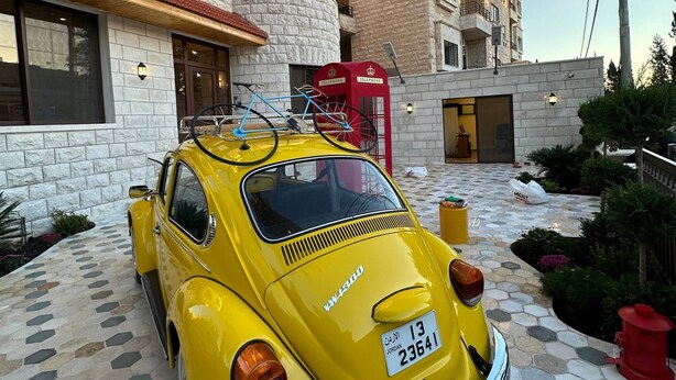 A vintage yellow car parked in front of a building with a stone facade and a red telephone booth. - 7 Antica Jabalal-lwebdeh (Amman)