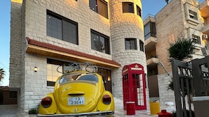 Un coche antiguo amarillo estacionado frente a un edificio de fachada de piedra, una cabina telefónica roja y una entrada con portones.