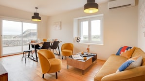 Una sala de estar moderna con un sofá, dos sillones amarillos, una mesa de centro de madera y una zona de comedor con mesa y sillas. Hay un ventanal con vistas al exterior.