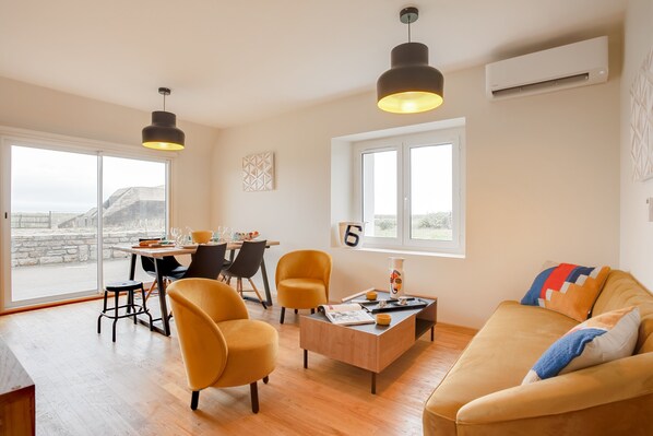 A modern living room with a sofa, two yellow chairs, a wooden coffee table, and a dining area with a table and chairs. There is a large window with a view of the outdoors.