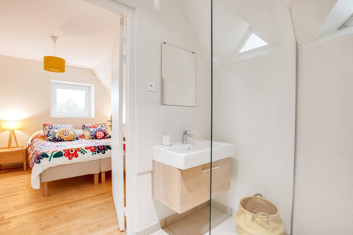 Une chambre à coucher moderne, avec un lit, une table de chevet, une lampe, et une salle de bain équipée d’un lavabo et d’un miroir.
