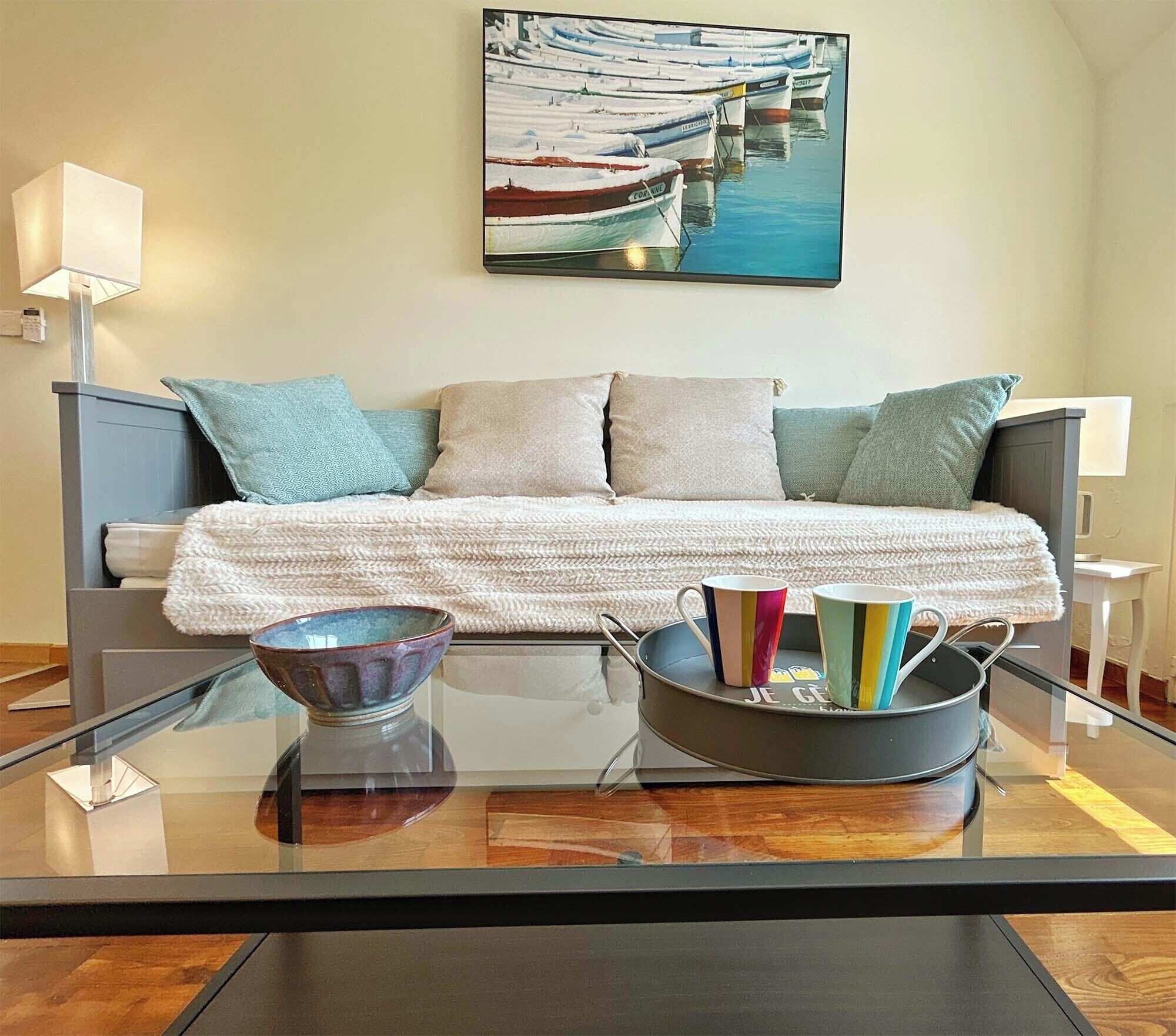Un salon moderne avec un canapé, des coussins, une table basse en verre et un tableau représentant des bateaux accroché au mur.
