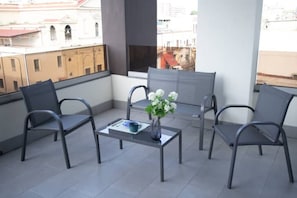 Un balcon avec quatre chaises en métal, une petite table ornée d’un vase de fleurs et une vue sur les immeubles, visible à travers une grande fenêtre.