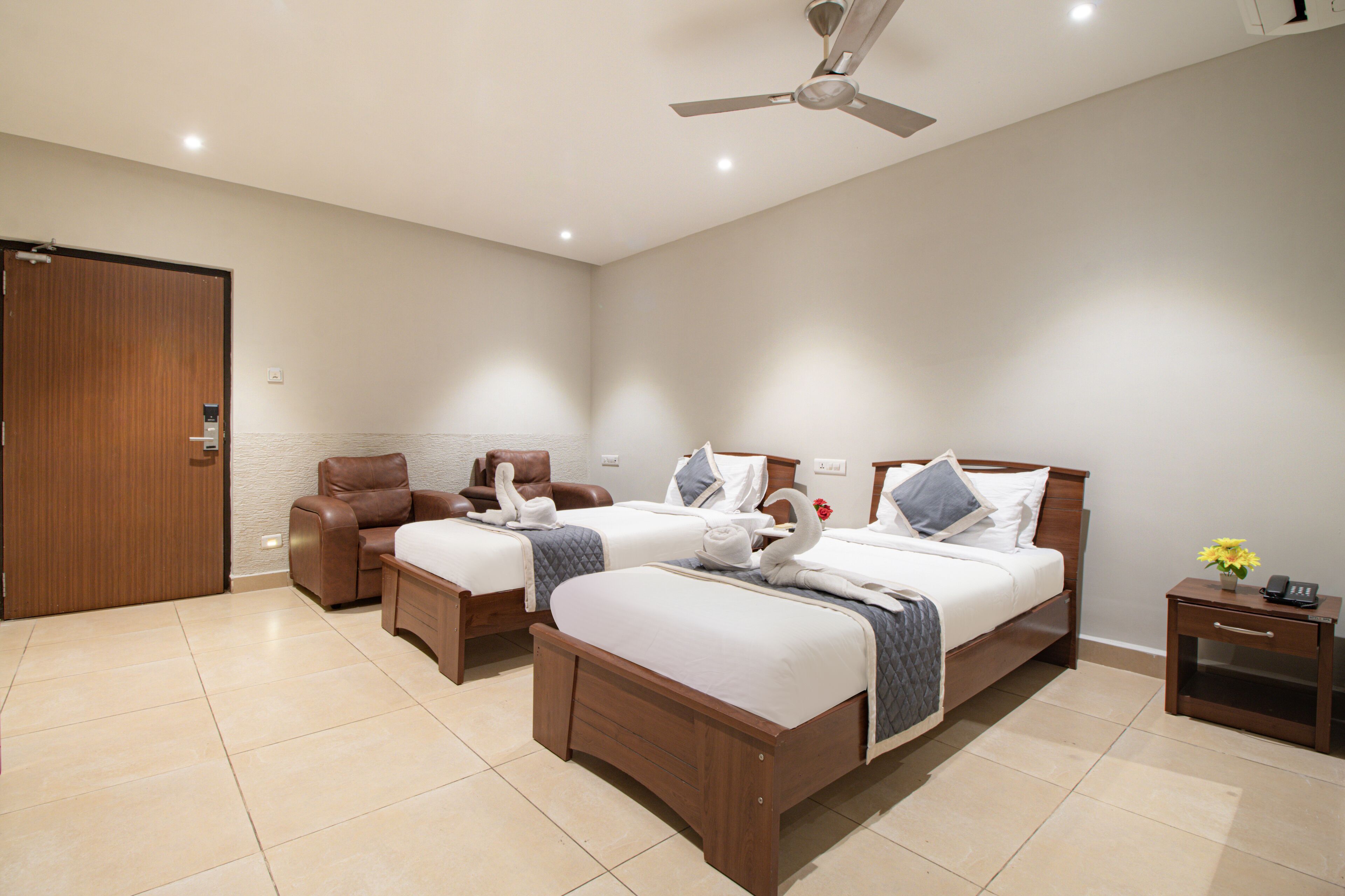 A hotel room with two beds, a wooden wardrobe, a leather armchair, a bedside table with a vase of flowers, and a ceiling fan.