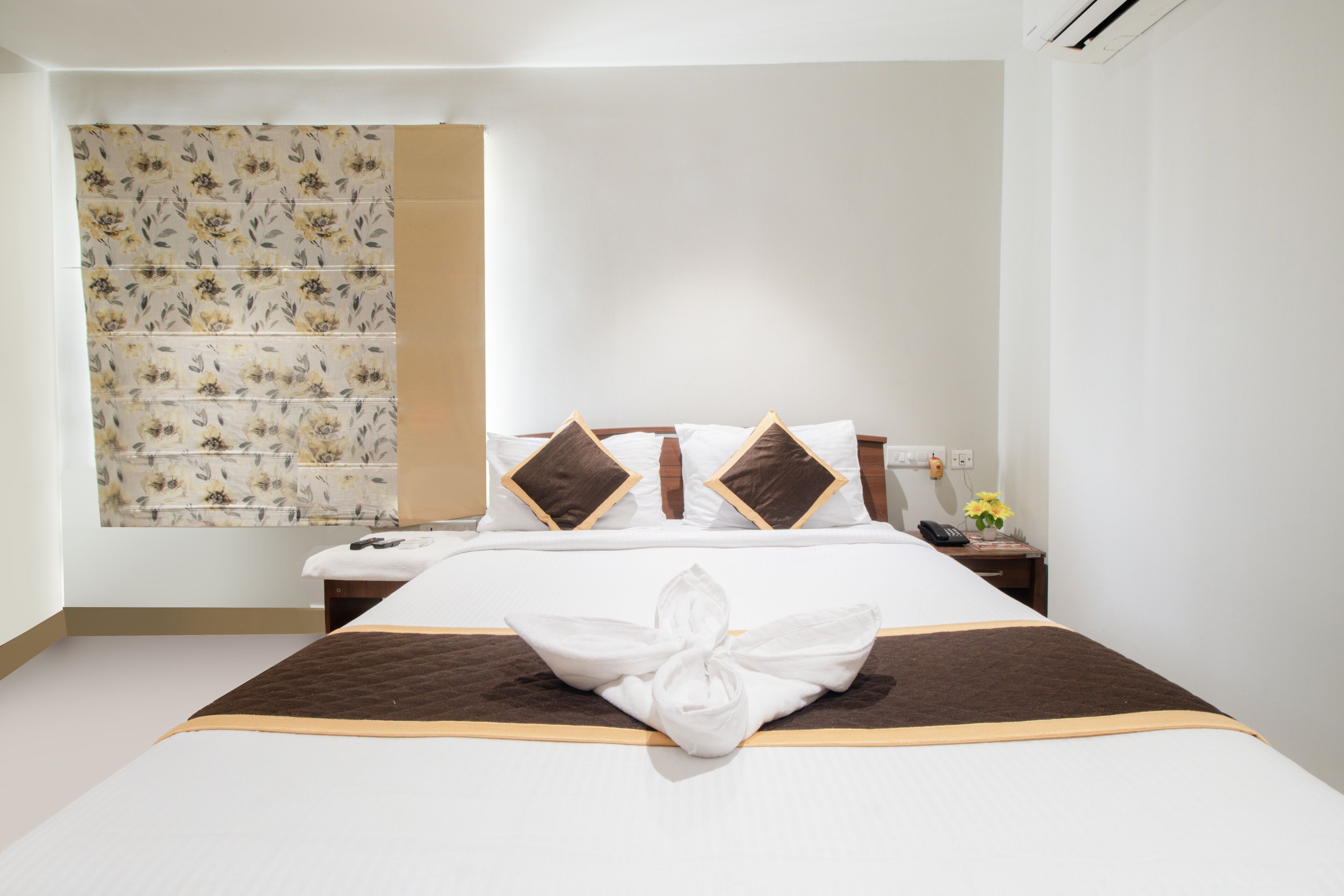A hotel room with a neatly made bed, a bedside table with a phone and a vase of flowers, and a wall with a floral-patterned curtain.