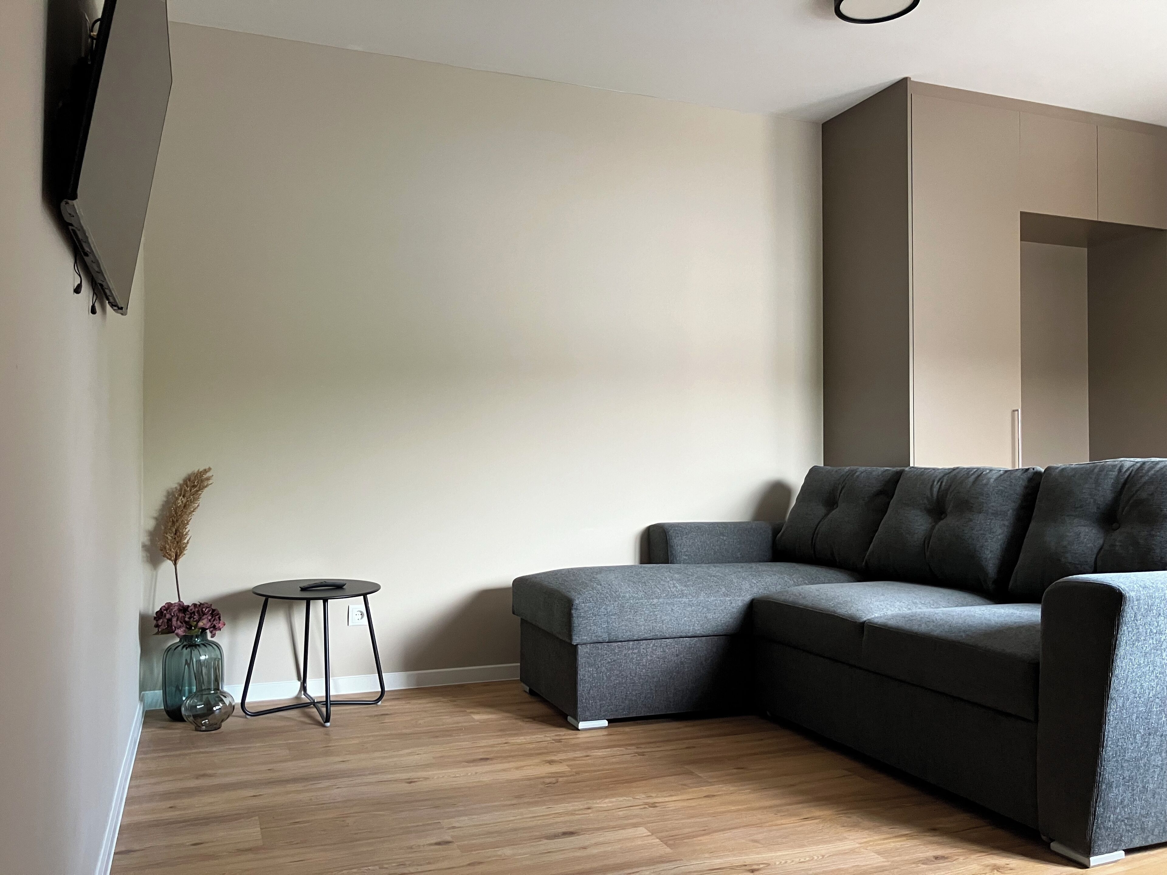 Ein modernes Wohnzimmer mit einem grauen Ecksofa, einem kleinen runden Tisch und einer Vase mit Trockenblumen.