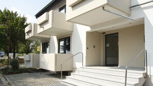 Un edificio moderno con techo plano, una escalera exterior que lleva a una puerta y un pequeño jardín con un árbol y un banco.