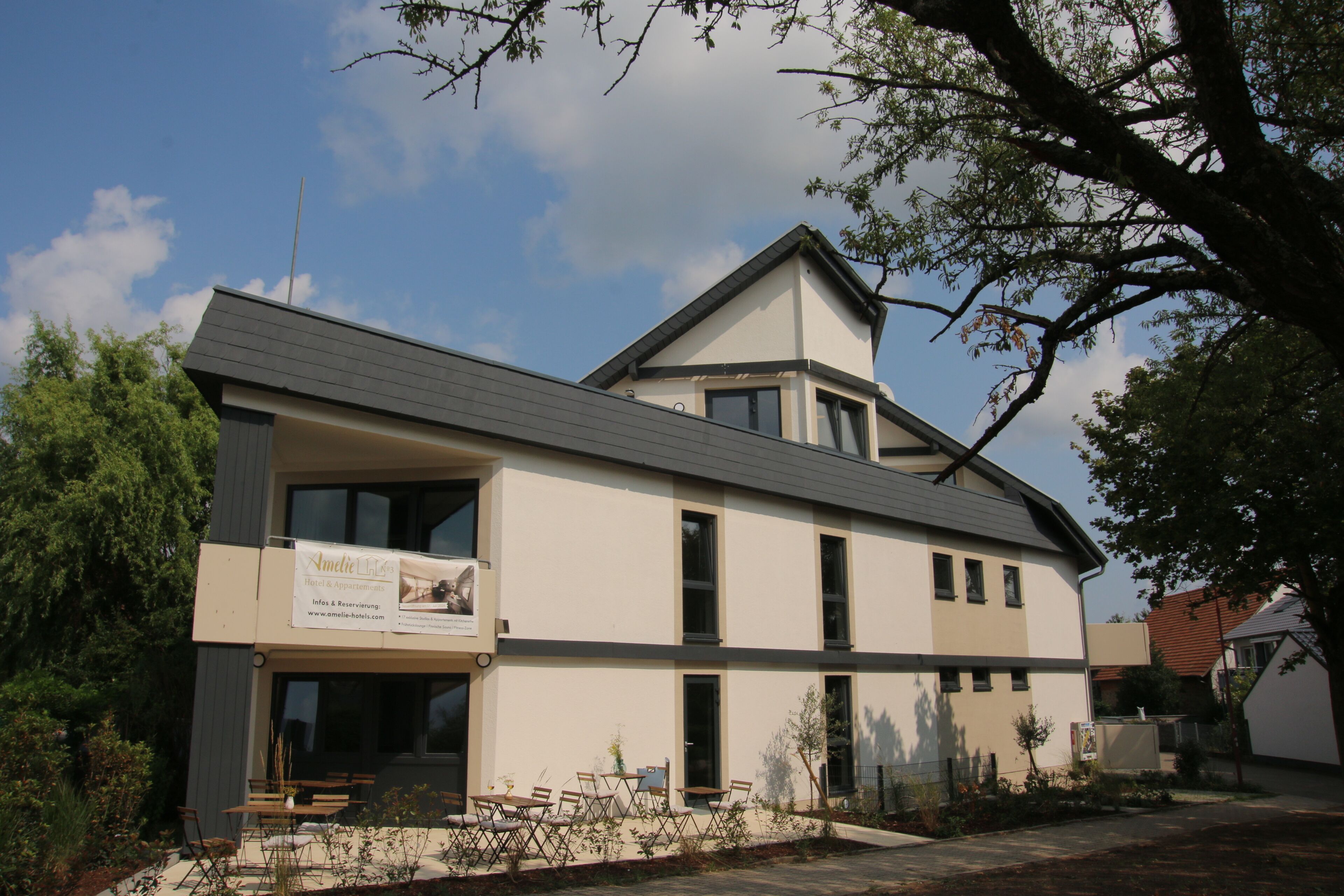 A modern two-story building with a flat roof and large windows. There is a sign on the building's facade, and outdoor seating is visible in front.