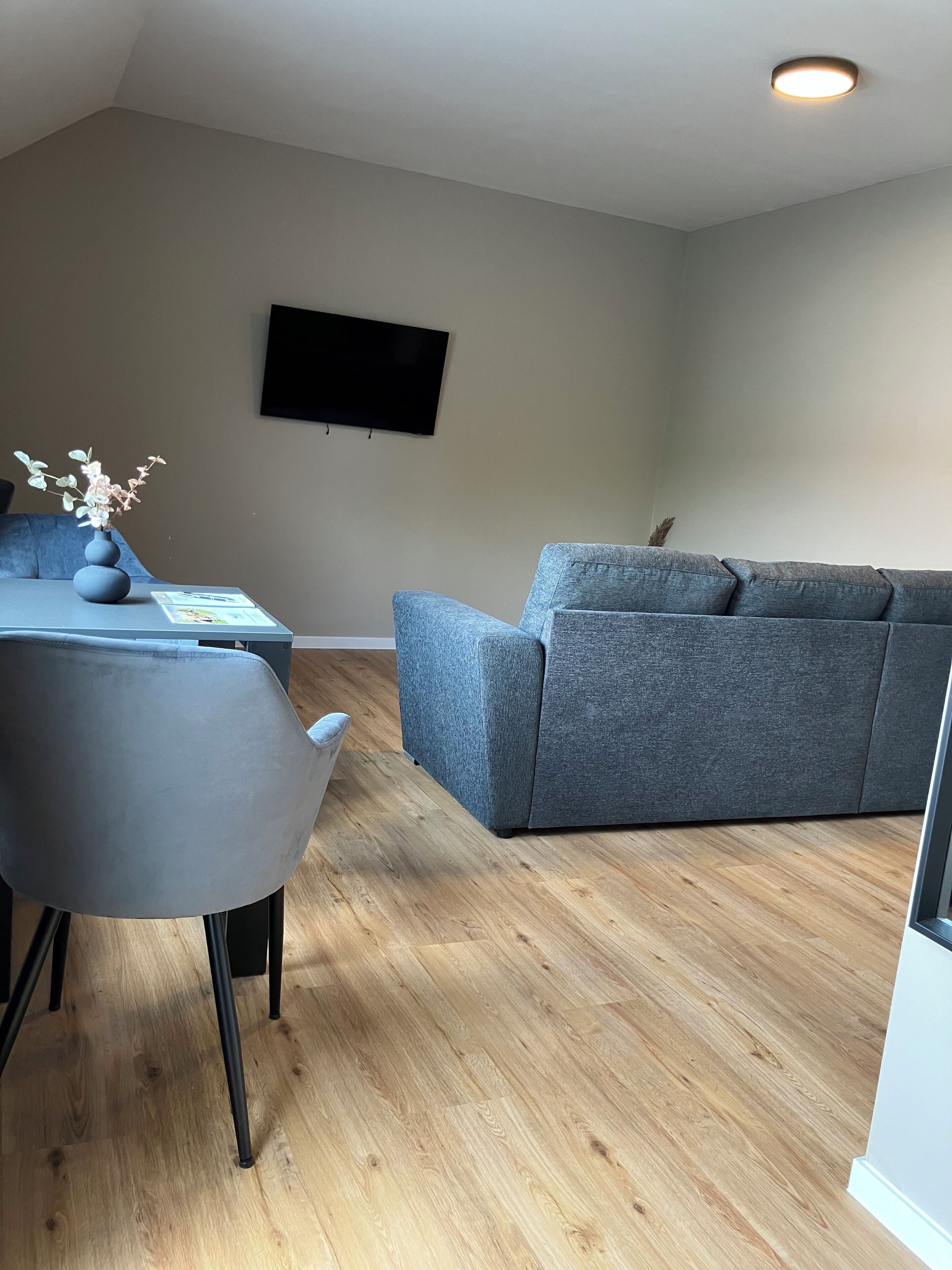 Una sala de estar moderna con un sofá y un sillón grises, piso de madera y un televisor de pantalla plana montado en la pared.