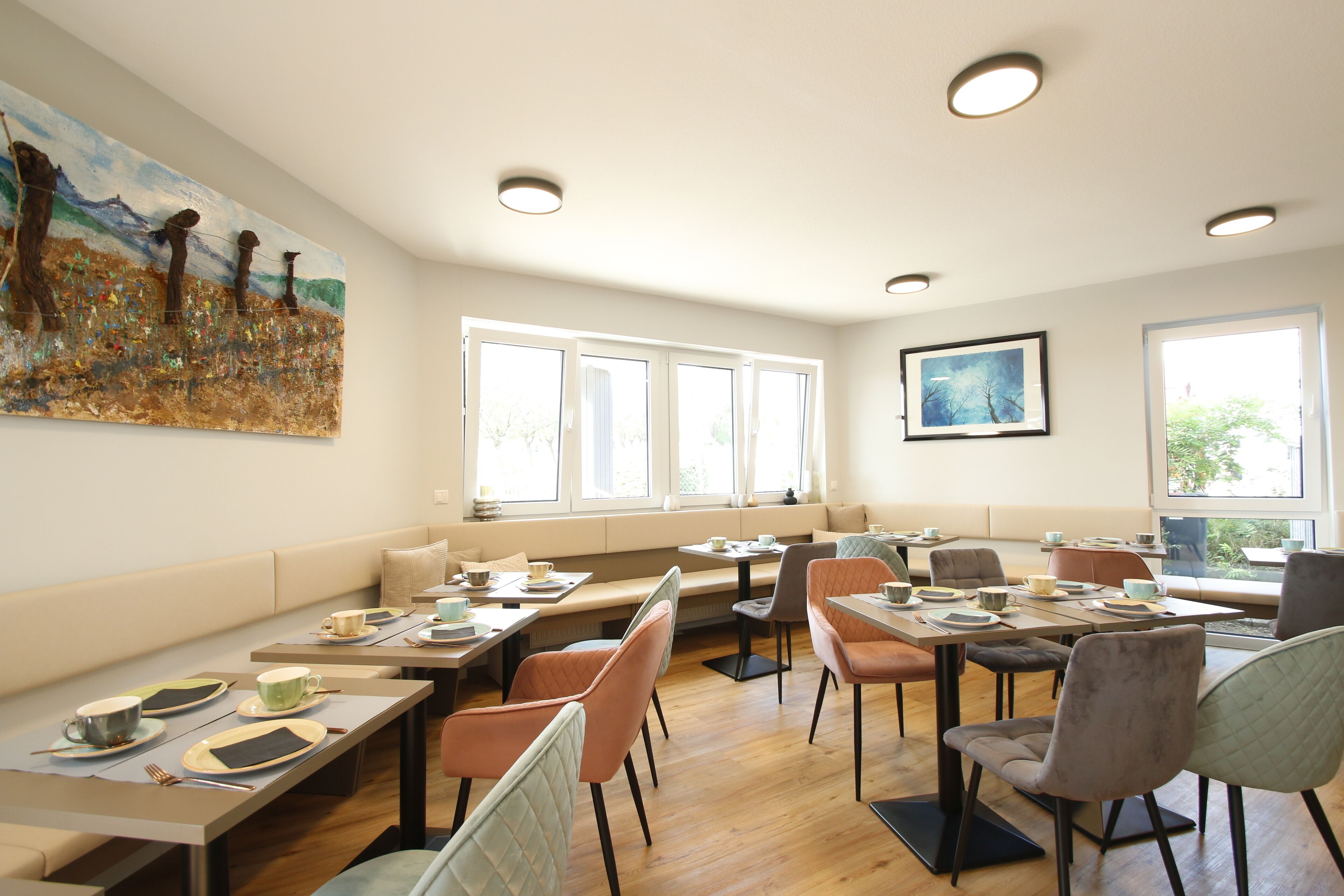 A dining area with tables and chairs, a large painting on the wall, and large windows allowing natural light.