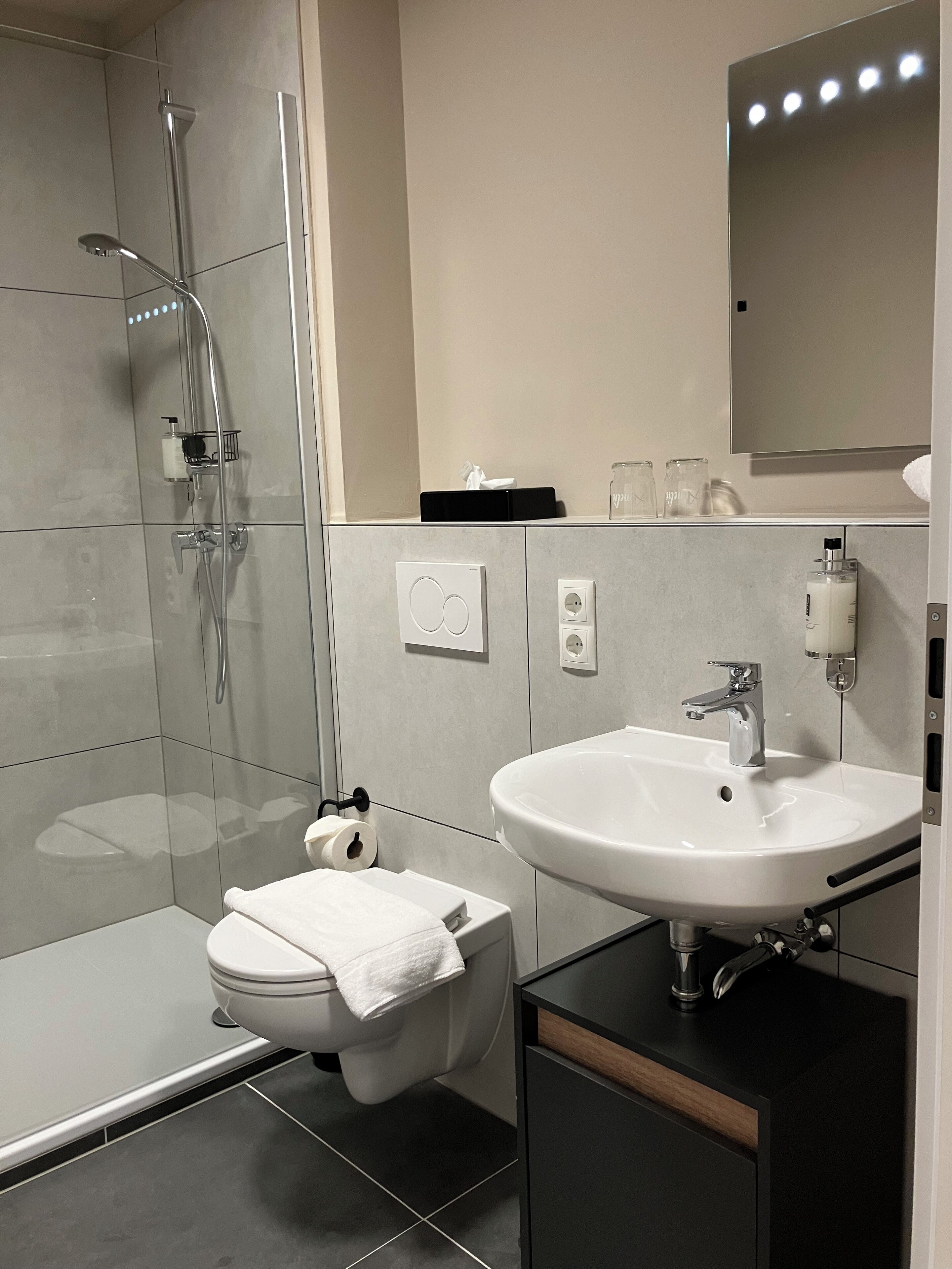 Une salle de bain moderne avec une douche à l’italienne en verre, des toilettes blanches, un lavabo avec un miroir au-dessus et un meuble noir en dessous.