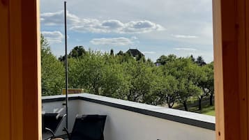 A balcony with two black lounge chairs and a table, overlooking a garden with trees and a clear sky.