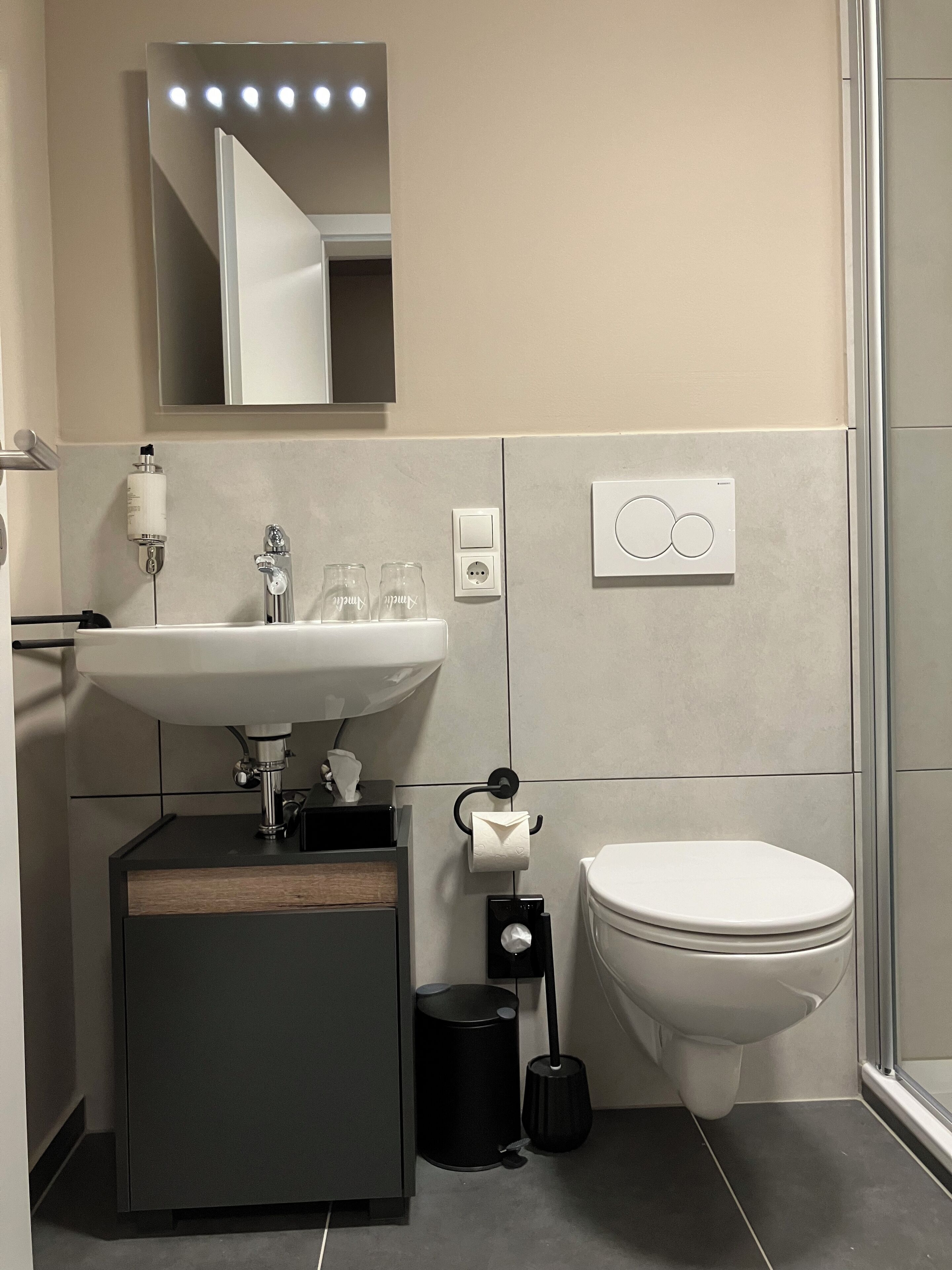 Ein modernes Badezimmer mit Waschbecken, Spiegel und Toilette in Weiß. Ein schwarzer Mülleimer und eine schwarze Toilettenbürste sind ebenfalls vorhanden.