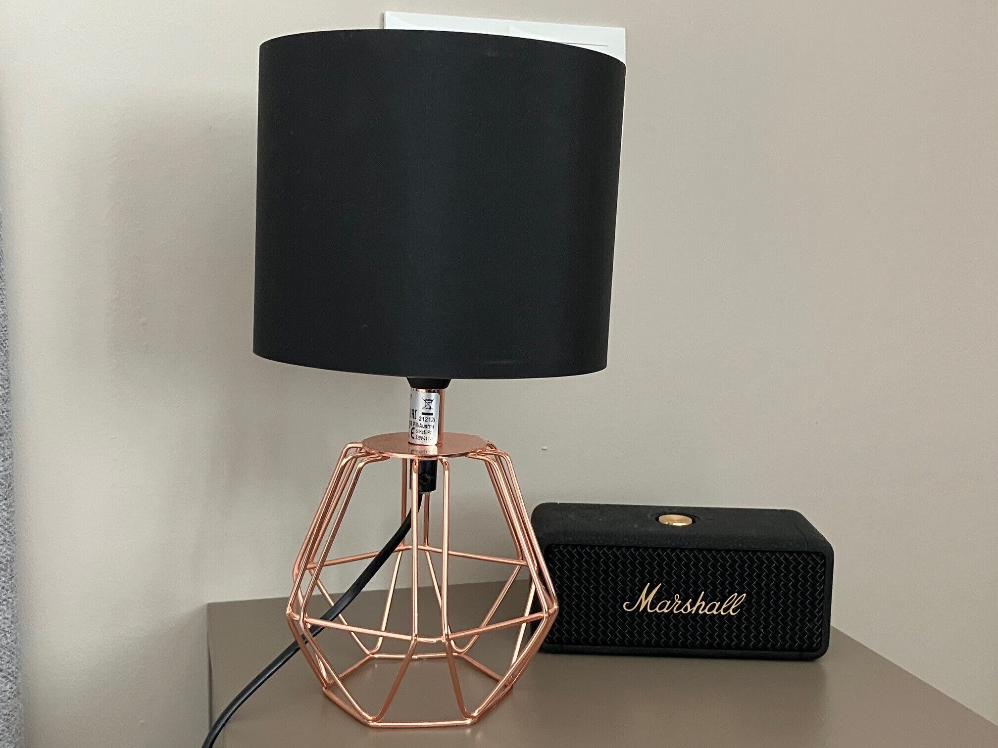 Eine Lampe mit schwarzem Lampenschirm und Kupferfuß, neben einem schwarzen Lautsprecher mit dem Aufdruck 'Marshall'.
