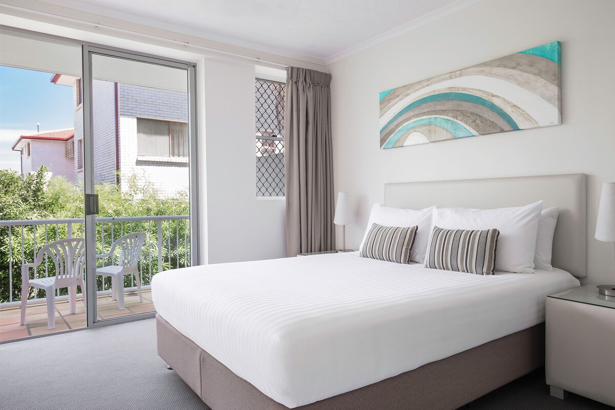A hotel room with a large bed, a balcony with chairs, and a view of buildings outside.