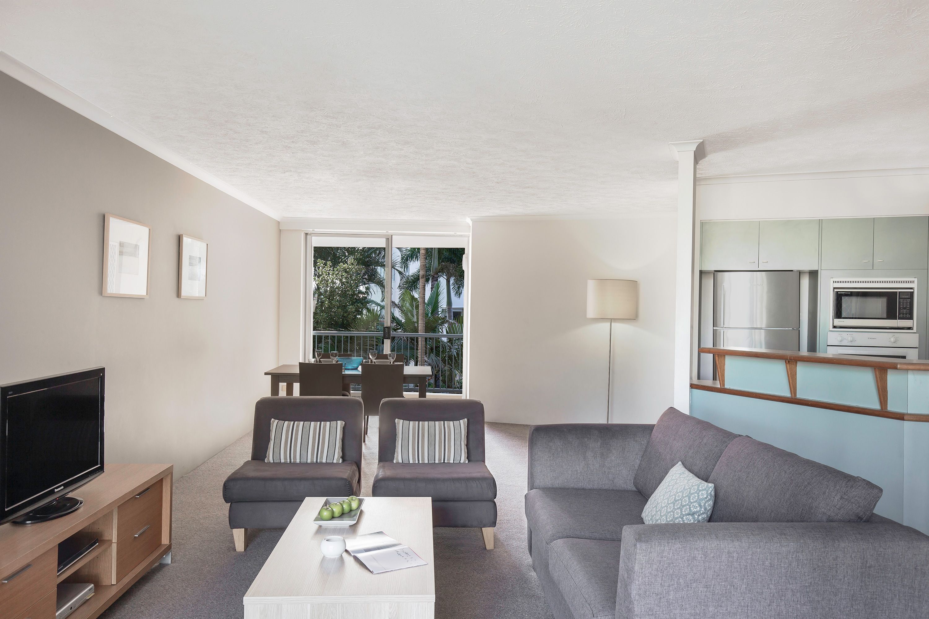 A modern living room with a flat-screen TV, grey sofas, a dining area with a table and chairs, and a kitchen with a microwave and refrigerator.
