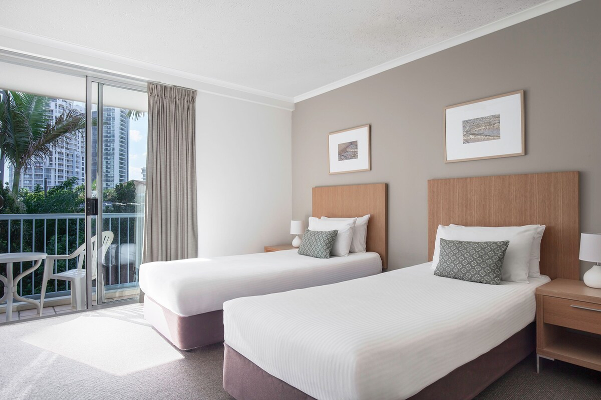 A hotel room with two beds, a balcony with a view of palm trees and buildings, and a small table with chairs.