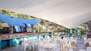 Zona de piscina al aire libre con un bar, sillas y mesas de plástico blanco.
