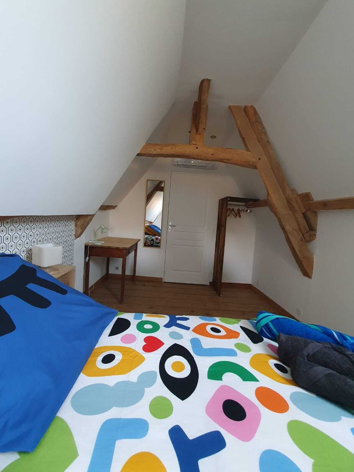 A small, cozy attic bedroom with a sloped ceiling, a wooden beam structure, a single bed with a colorful bedspread, a small desk, and a door leading to another room.