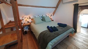A bedroom with a loft bed, wooden beams, a ladder, a bedside table with a lamp, and a window with curtains.