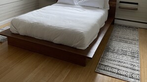 A bedroom with a wooden bed, white bedding, a patterned rug, a window with blinds, and a sloped ceiling.