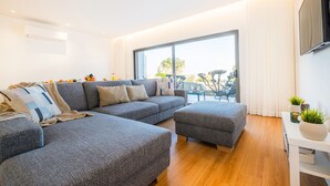 Een moderne woonkamer met een grijze modulaire bank, een klein voetenbankje en een witte salontafel. Er is een groot raam met gordijnen, een flatscreen televisie aan de muur en een kleine tafel met planten.