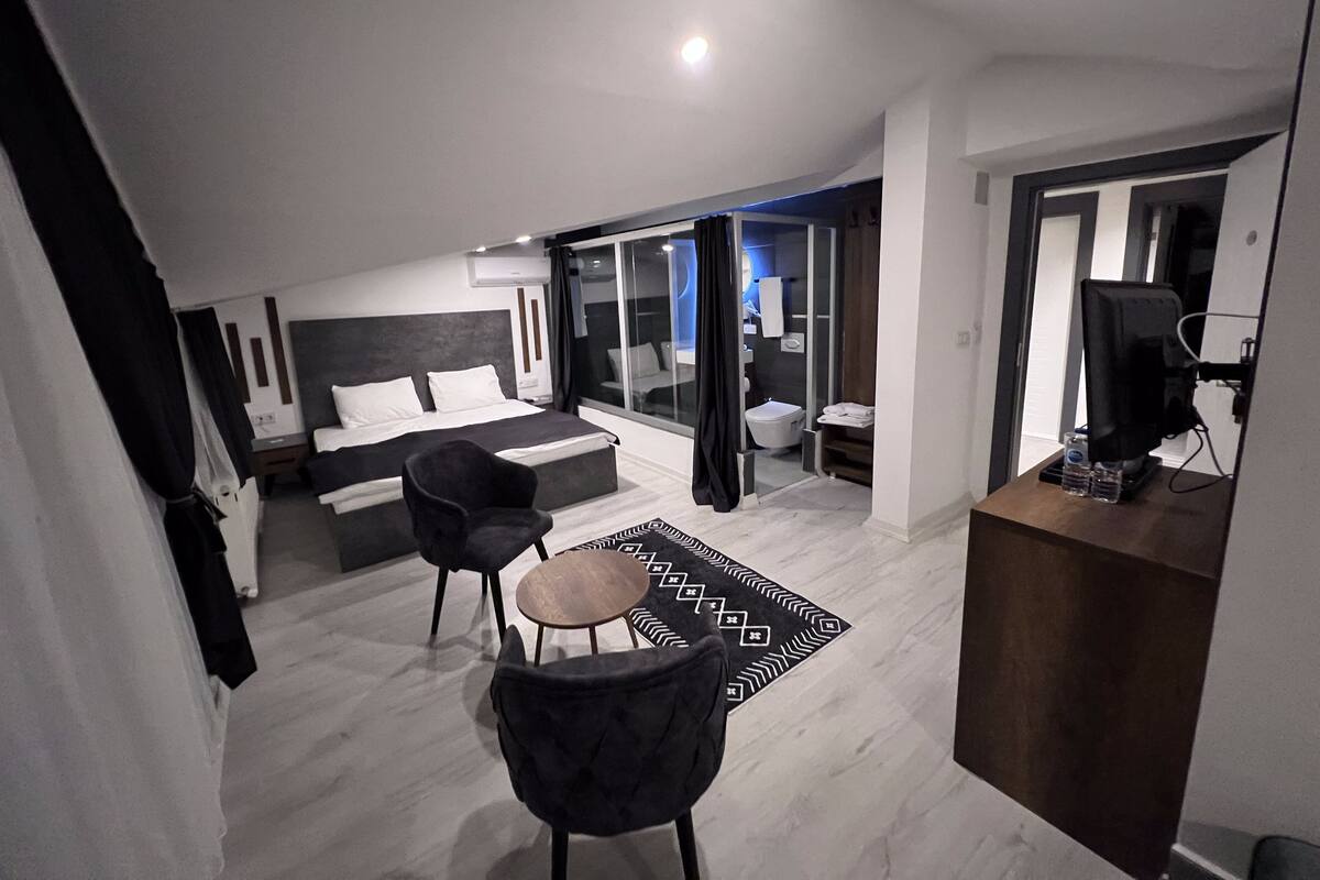 A modern hotel room with a large bed, a desk, a chair, and a bathroom visible through a glass door.