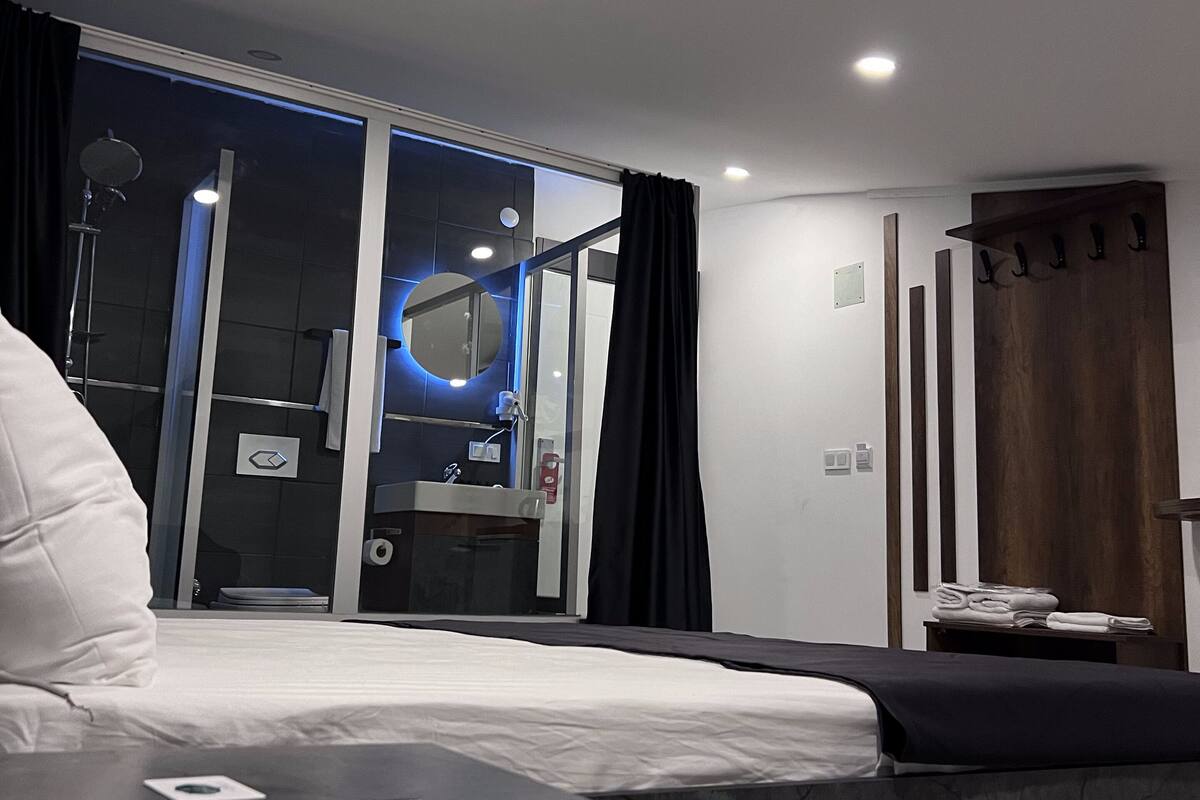 A modern hotel room with a bed, a bedside table, a bathroom visible through a glass door, and a wooden coat rack.