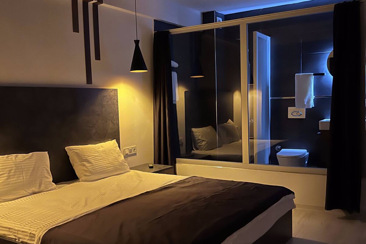A modern bedroom with a large bed, bedside tables, a mirror, and a bathroom visible through a glass door.