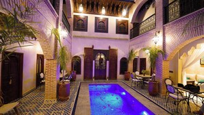 Un patio con piscina, suelo de baldosas y arquitectura marroquí tradicional, que incluye arcos y puertas de madera.