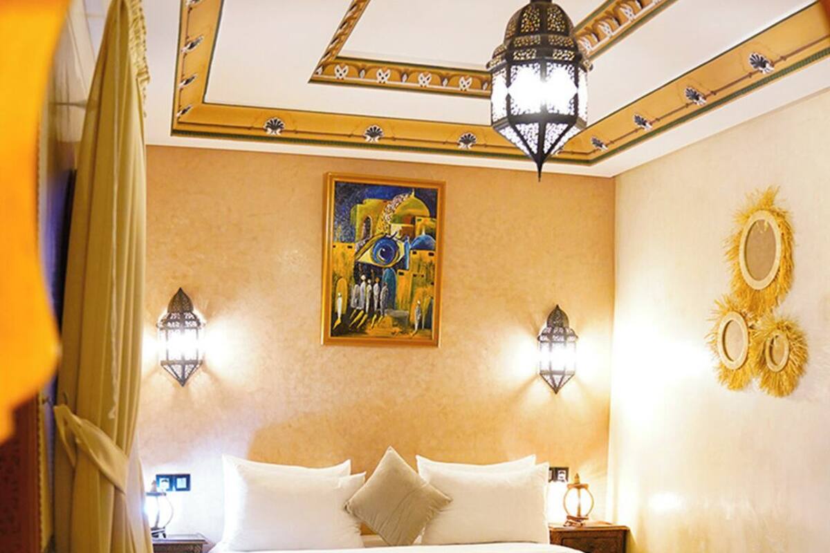 Habitación de hotel con cama, dos lámparas de pared, un cuadro, un espejo decorativo y un tapete estampado.