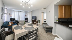 A modern living room with a dining table, sofa, and TV. There is a kitchen area with wooden cabinets and a countertop.