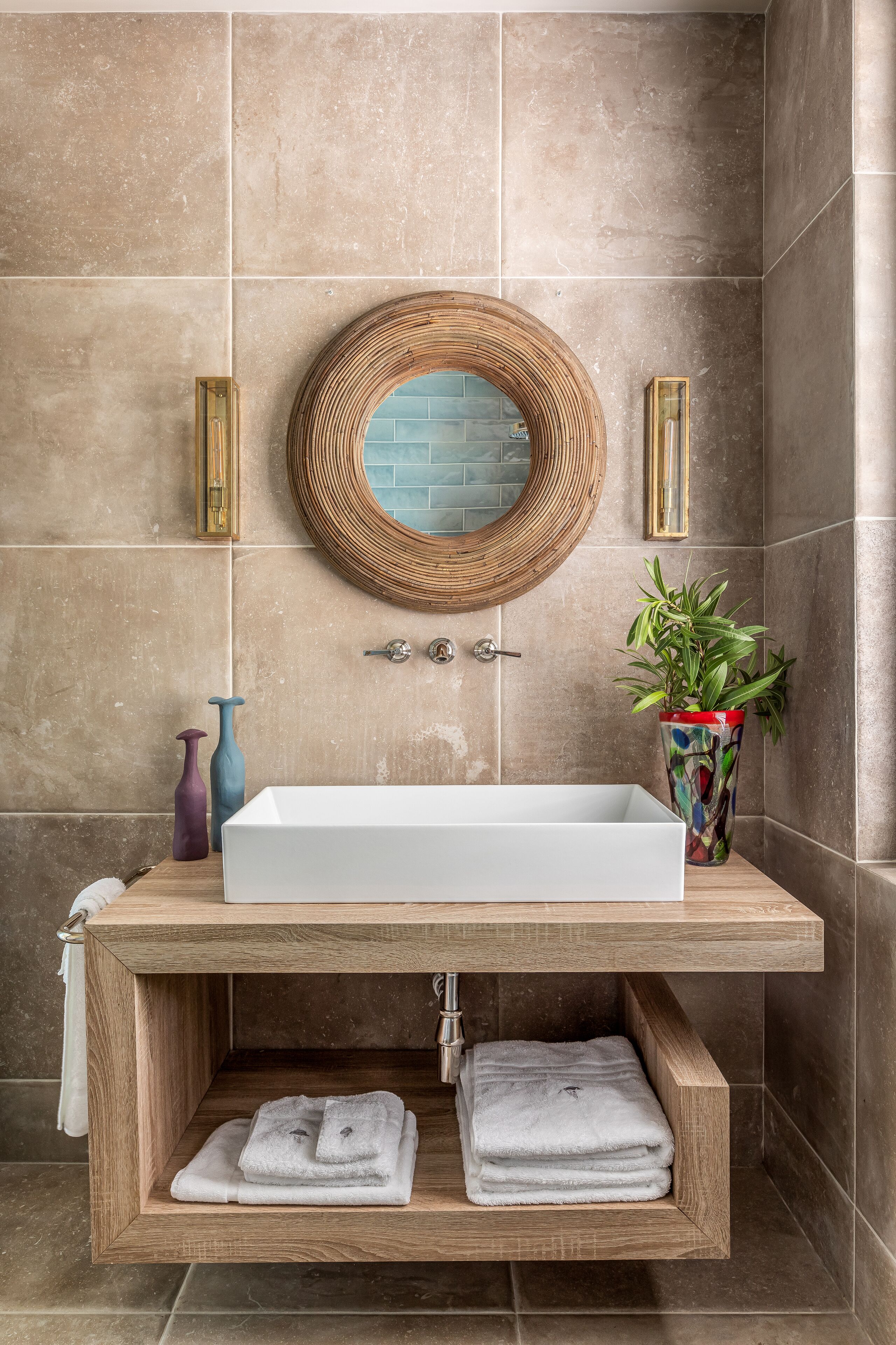 A bathroom with a rectangular sink, a round mirror, a potted plant, and a wooden shelf with towels.