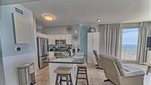 A modern kitchen with a central island, white cabinets, stainless steel appliances, and a view of the ocean through a large window. - Shores of Panama 503~End Unit 3 Bdrm (Panama City Beach)