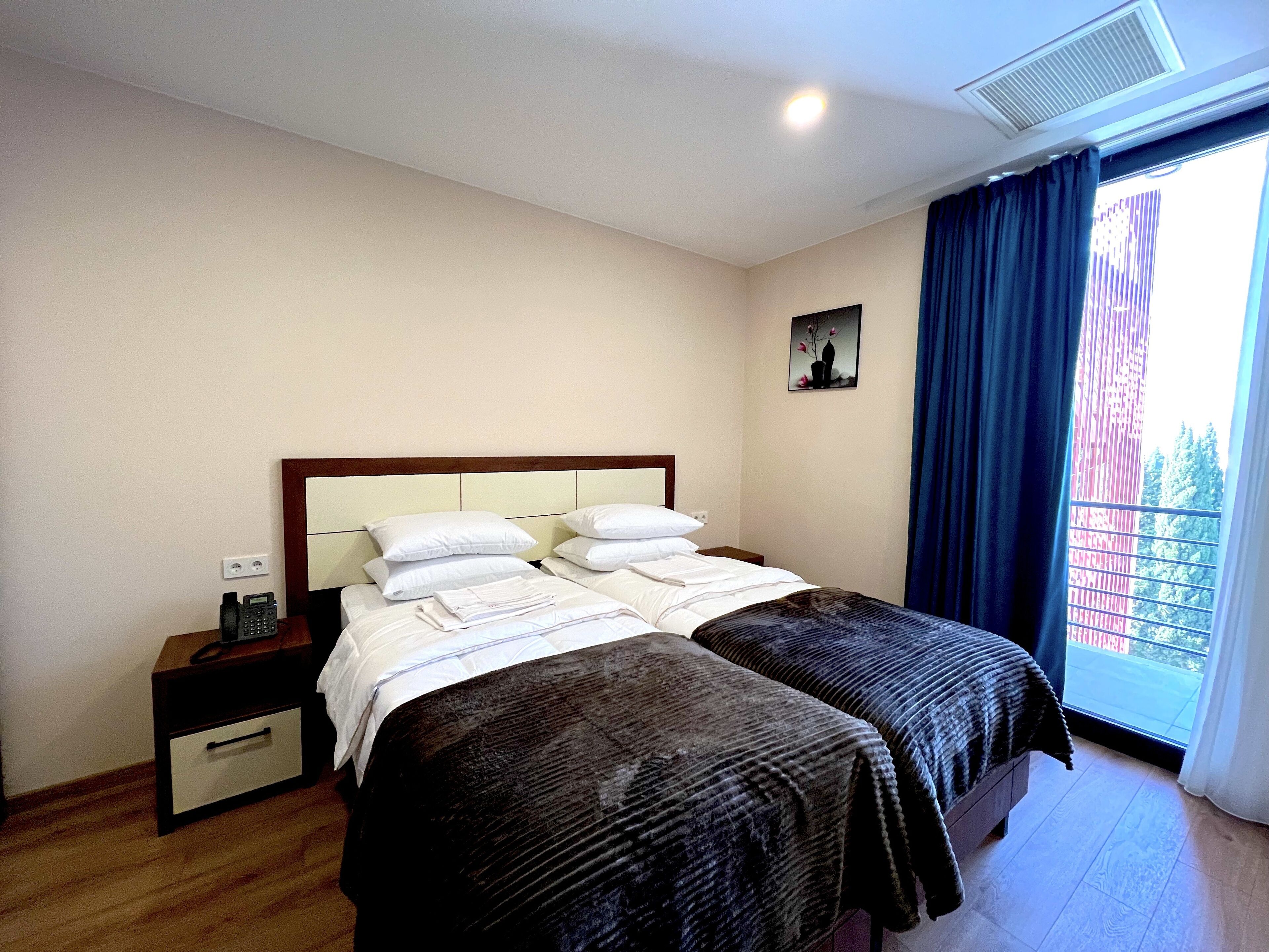 A hotel room with a double bed, a bedside table, a small drawer unit, a painting on the wall, and a window with curtains.