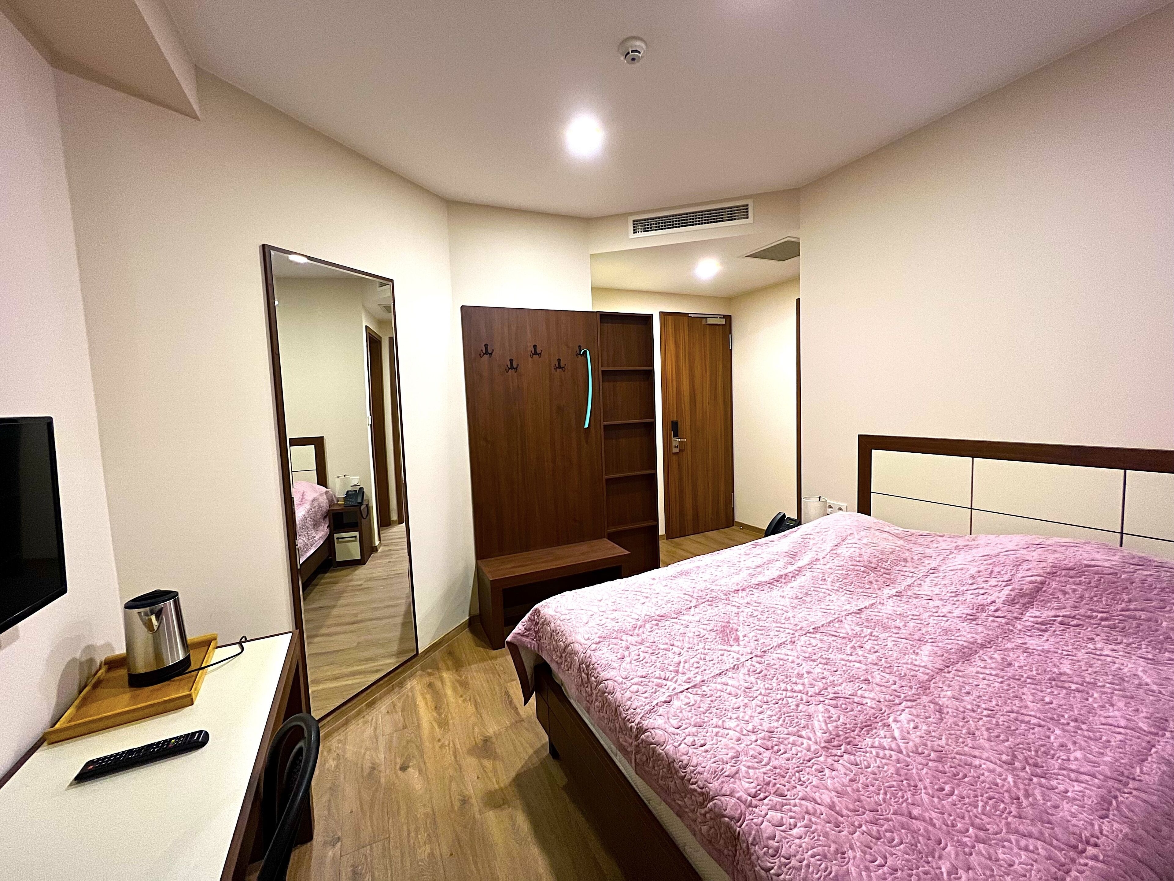 A hotel room with a bed, a desk with a kettle, a remote control, a wardrobe, and a mirror.