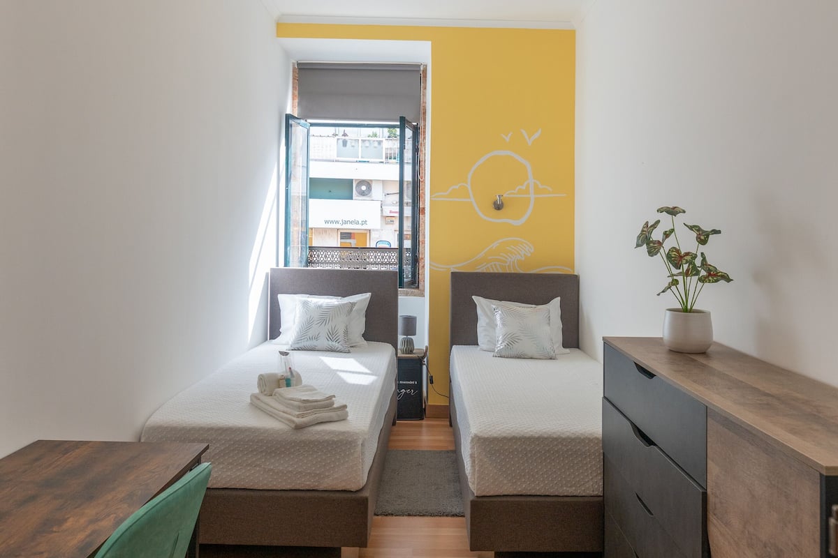 A modern hotel room with two beds, a wooden desk, a green chair, and a view of the city through a window.