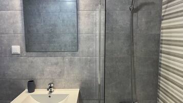 Een badkamer met een wit wastafel, een spiegel, een douche met een handdouche en grijze betegelde muren.