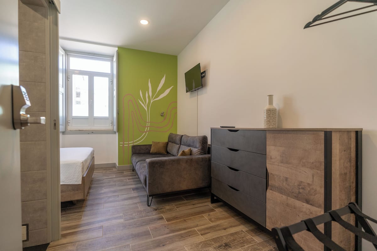 A bedroom with a bed, a green accent wall, a grey sofa, a wooden dresser, and a TV mounted on the wall.