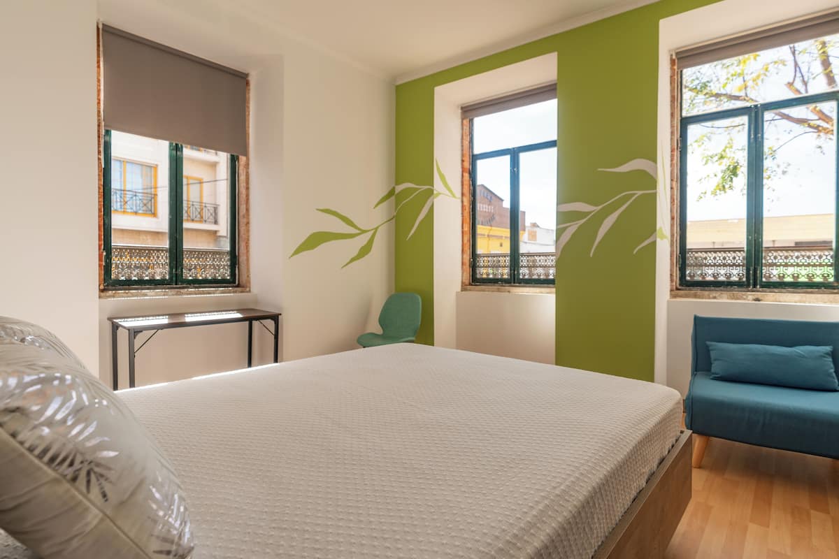A bedroom with a large bed, a green accent wall, a blue sofa, and a window with a view of buildings.