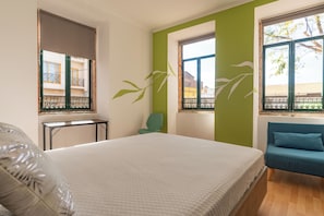 Quarto com cama grande, parede verde, sofá azul e janela com vista para edifícios.