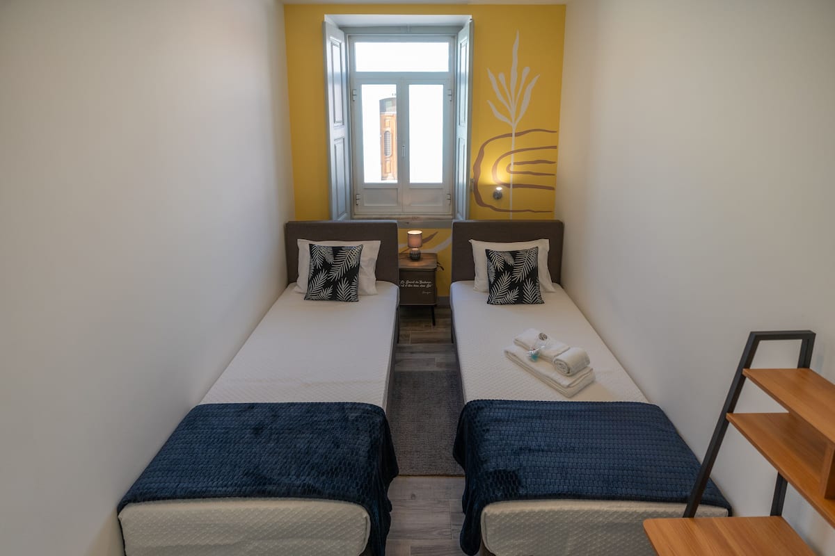 A small, bright room with two single beds, a small table with a lamp, and a window with a view of a building.