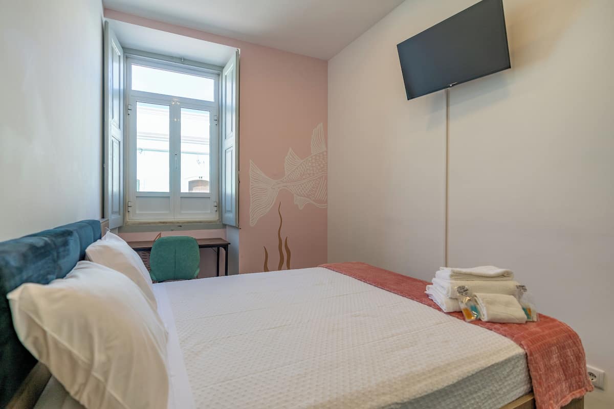 A hotel room with a bed, a desk, a chair, a TV mounted on the wall, and a window with a view of the outside.