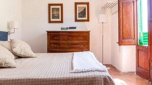 A bedroom with a bed, a wooden dresser, two framed pictures on the wall, and a window with a green view. - Villa La Fenice by Wonderful Italy (Castellammare del Golfo)