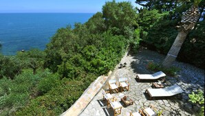 A stone patio with lounge chairs overlooking a body of water, surrounded by dense greenery. - Villa La Fenice by Wonderful Italy (Castellammare del Golfo)