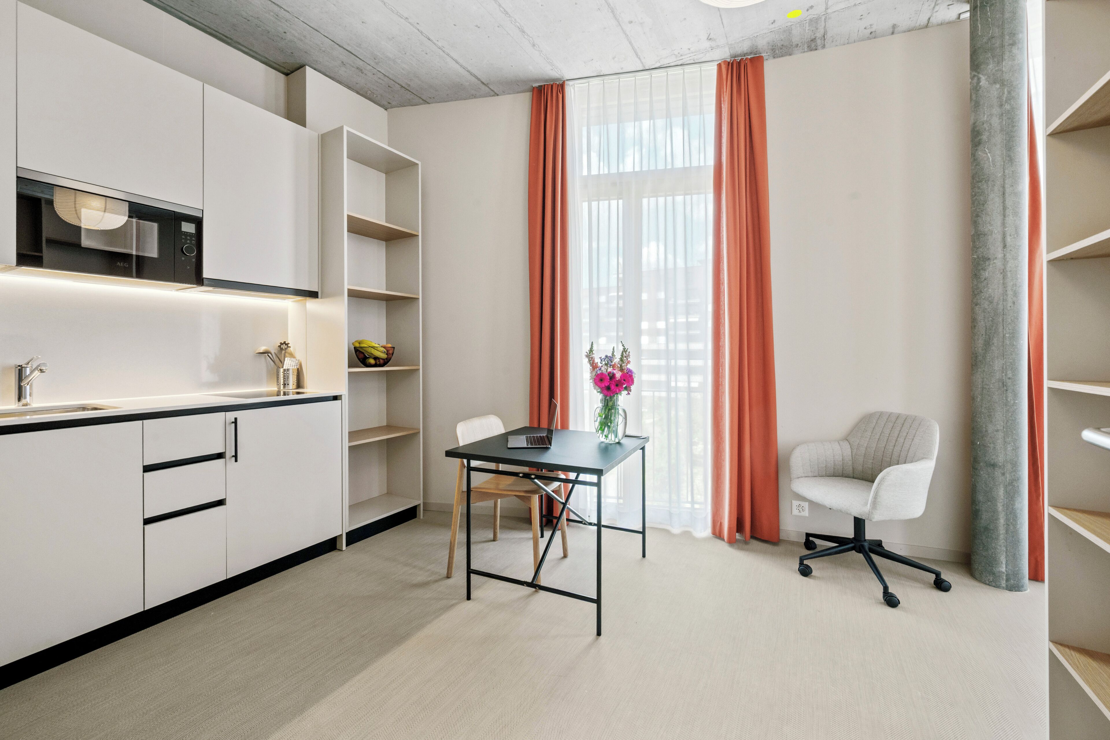 Una cocina moderna con armarios blancos, encimera negra y microondas integrado. Hay una mesa de comedor con una computadora portátil y un florero. Cortinas naranjas enmarcan una ventana, y se observa una silla de oficina blanca.