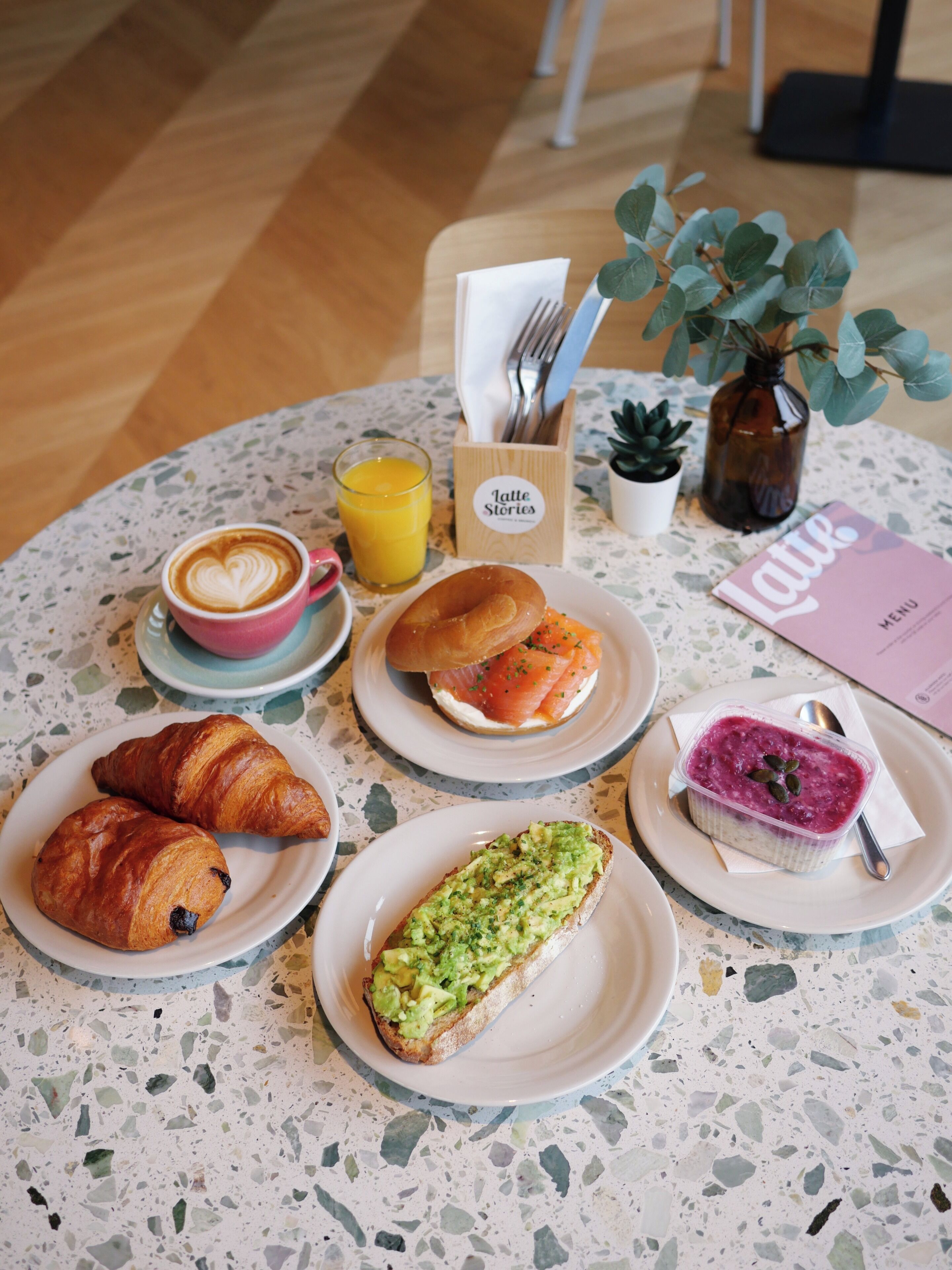 Ein runder Tisch mit einer Auswahl an Frühstücksleckereien, darunter Croissants, ein Smoothie, ein Croissant mit Avocado, ein Croissant mit Schokolade, ein Croissant mit Marmelade, ein Croissant mit latte-art-Herzchen und ein Croissant mit Blaubeeren.