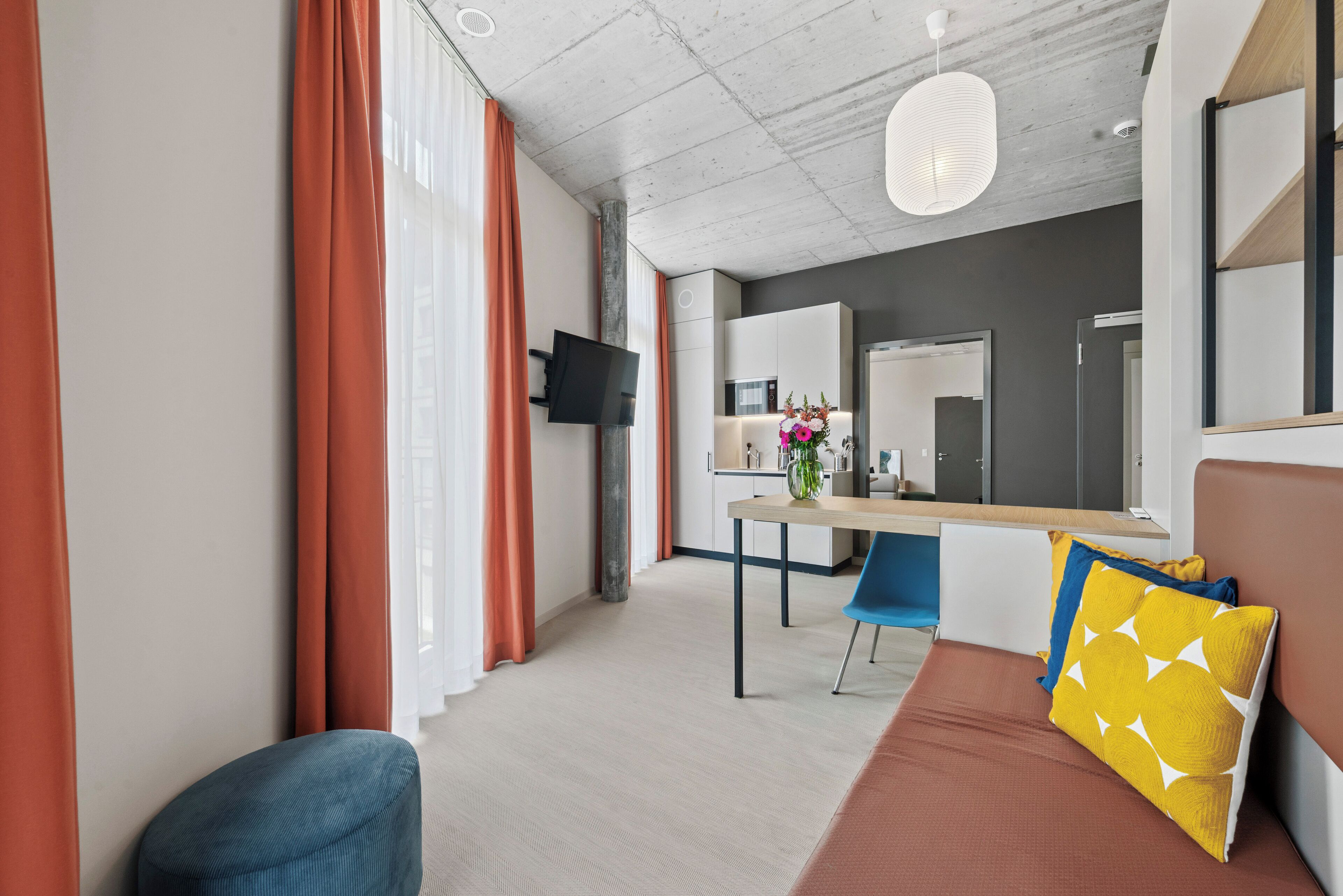 Une chambre d’hôtel moderne dotée d’un coin repas, d’un canapé avec des coussins, d’un téléviseur fixé au mur et d’un grand miroir.