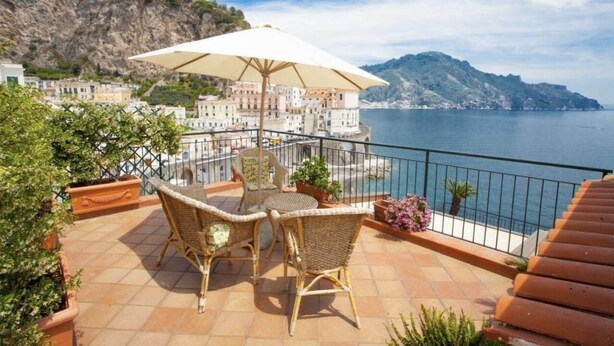 Eine Terrasse mit Korbstühlen und einem Tisch unter einem Sonnenschirm, mit Blick auf eine Küstenstadt und das Meer.